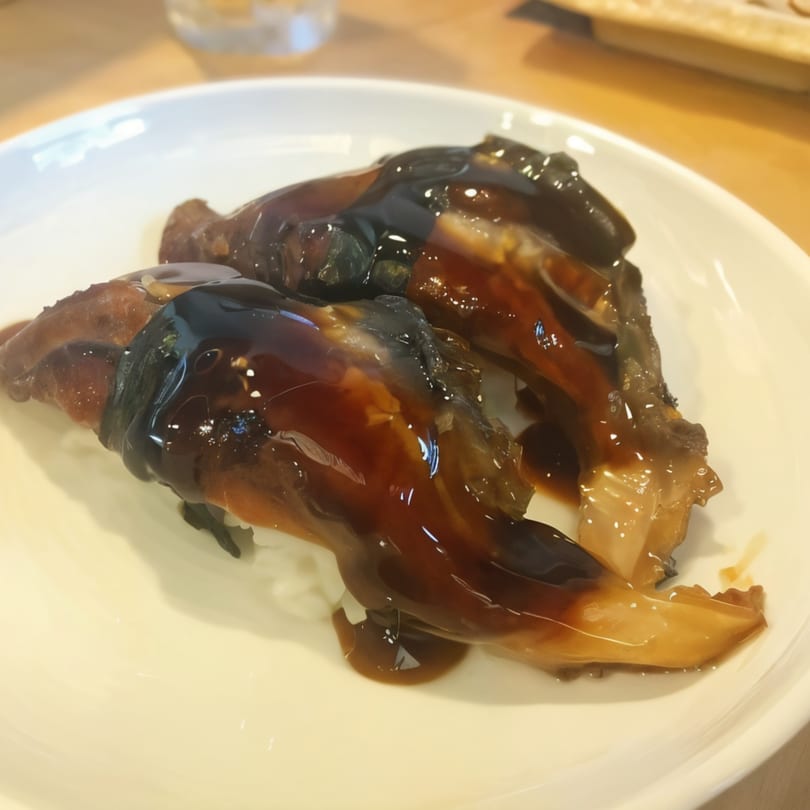 Two pieces of dark, glossy eel sushi with sweet sauce on a white plate at Sushi Bei.