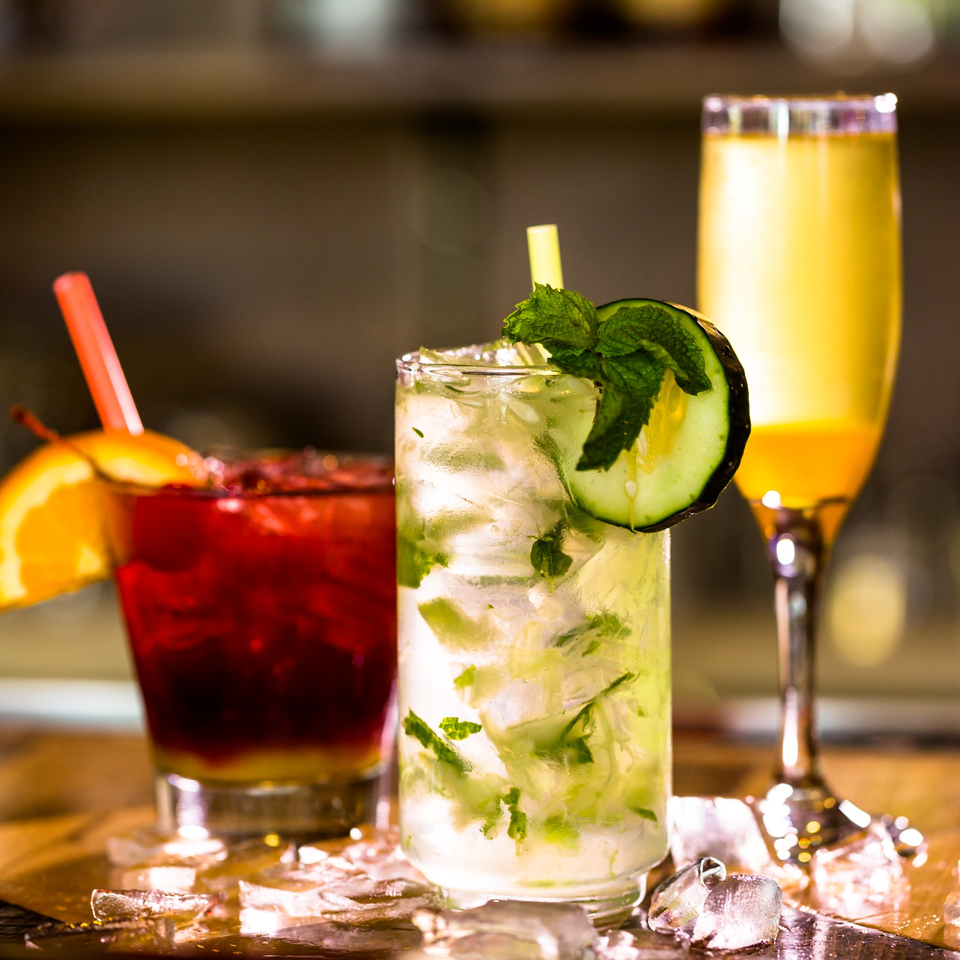 Three vibrant cocktails, including a mojito with cucumber and mint, on a bar with ice.